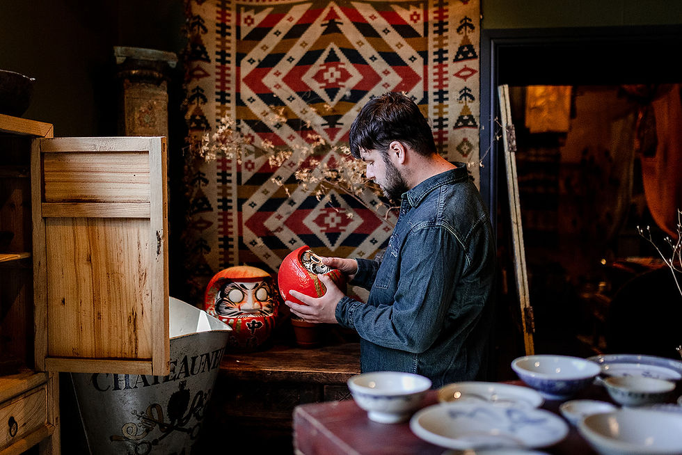 Man in a denim shirt examines a red daruma doll in a cozy shop, with patterned tapestry, wooden shelves, and ceramic bowls nearby, small business branding photography.