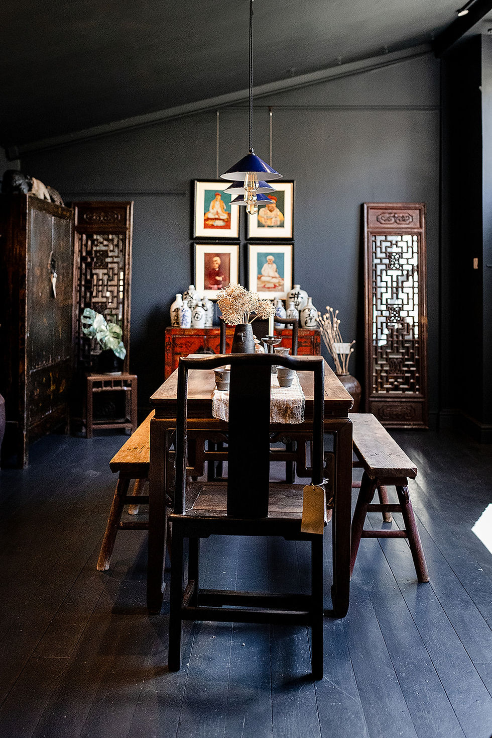 Dim vintage dining room with dark wood table, bench, black chair, hanging lamp, framed art, and ceramic vases on a cabinet