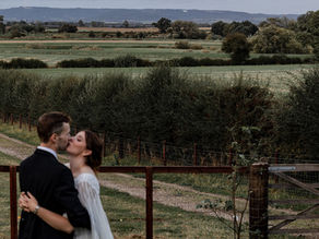Newlyweds kiss beside a wooden gate in a green countryside field with rolling hills and trees. At Lodge Farm Cundal