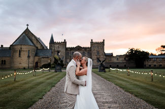 Sneaton Castle Wedding couple kiss with pink sunset and castle in the background there is festoon lighting leading towards the castle and moody romantic feeling.