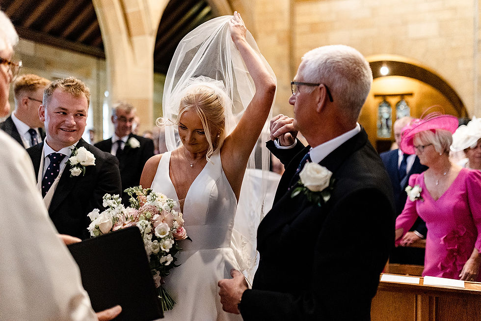 Carlton-in-Cleveland wedding. Bride lifts veil in church as guests watch; groom smiles beside her holding bouquet, with a woman in bright pink hat nearby.