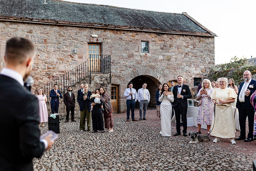 Wedding guests toast in a stone courtyard; bride and groom stand smiling, with a small dog on a leash in the foreground. relaxed Cumbria wedding photography