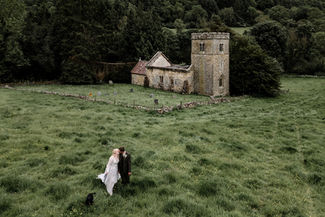Bride and groom walking hand in hand through long grass with their black dog, in front of a decommissioned stone church surrounded by trees, North Yorkshire countryside.