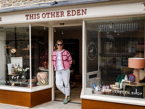 Woman in pink jacket stands at the doorway of This Other Eden shop, with garden and homeware displays in the windows.