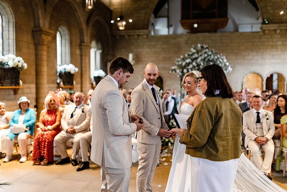 Bride and groom exchange rings during a wedding ceremony in a stone church, with guests seated behind them and a smiling officiant