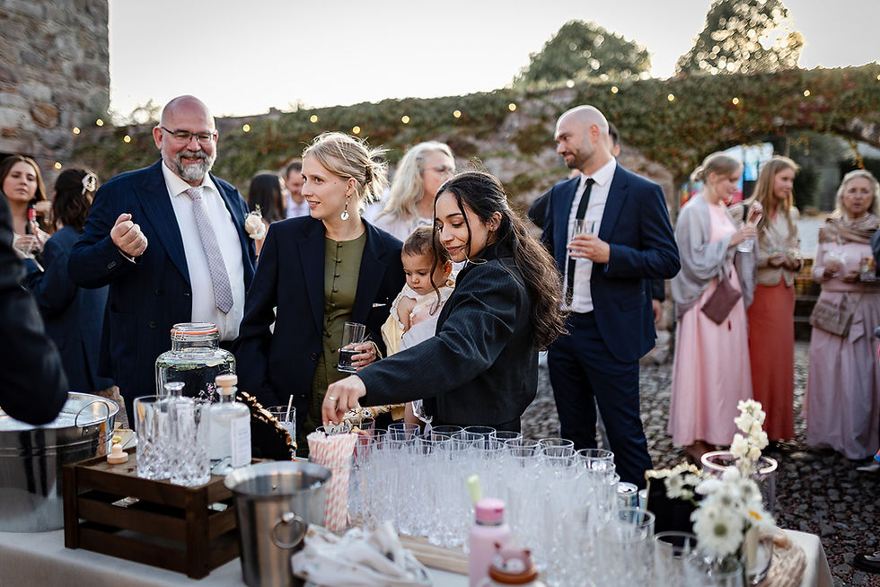 Guests mingle at an outdoor reception table, pouring drinks; a woman holds a baby, with stone walls and string lights behind them. relaxed Cumbria wedding photography