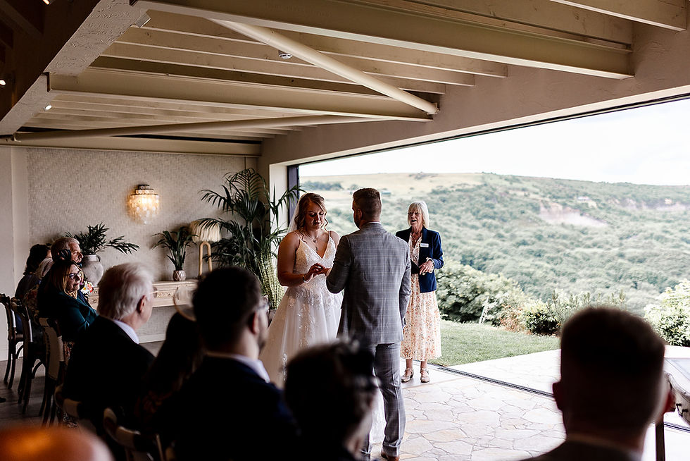 Bride and groom exchange rings at an indoor wedding ceremony overlooking green hills, with guests seated nearby.