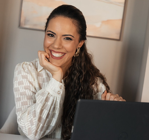Mulher sorridente com a mão no queixo sentada em frente a um notebook.