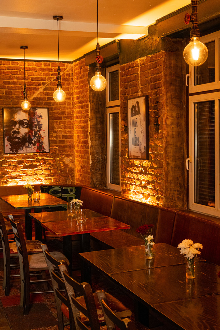 Warmly lit dining area at Bariton restaurant featuring exposed brick walls, pendant lights, cozy wooden tables, and artistic decor, including portraits and ‘War is Over’ artwork.