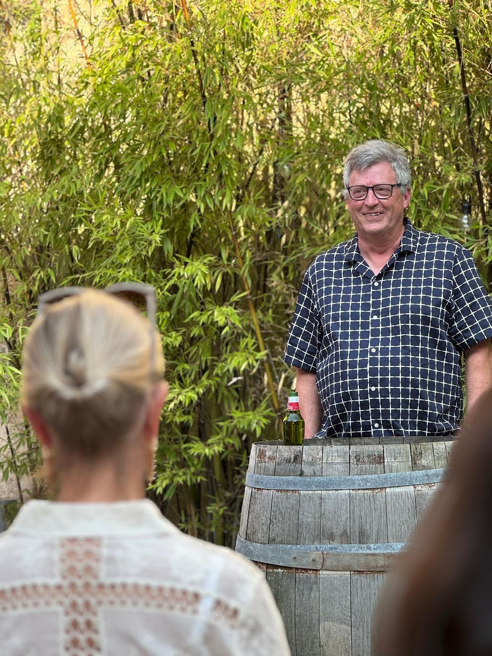 Man, stood at barrel facing, with happy appearance giving a speech to a group of people,  The background is a courtyard with bamboo in the background.