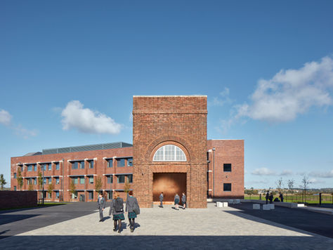Students in uniforms walk near a modern red brick school building under a clear blue sky. Playing fields are visible in the background.
