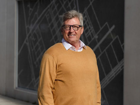 Smiling person in a tan jumper stands in front of a dark wall with a geometric pattern. Bright demeanor, urban setting.