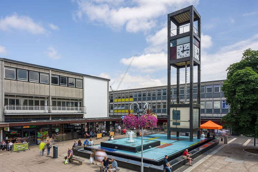 Stevenage's town square with tall clock tower, fountain, seated pedestrians, and shops including Starbucks and Subway under a blue sky
