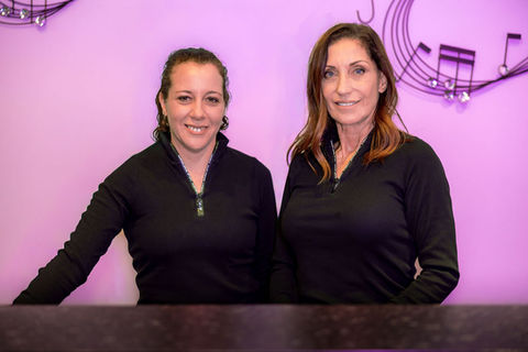 Two women smiling in black zip-up tops against a purple wall.