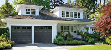Modern white two-story home with dark gray carriage-style double garage doors, large front windows, landscaped shrubs and flowers, and a clean concrete driveway surrounded by mature trees.