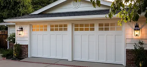 Traditional white garage doors with upper grid-style windows on a craftsman-style home, featuring black exterior lantern lighting, brick accents, and a clean concrete driveway surrounded by landscaping.