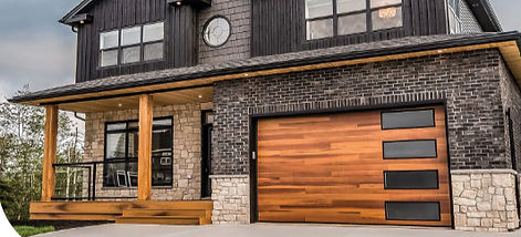 Modern two-story home with dark siding, stone accents, and a contemporary wood-tone garage door featuring sleek black glass windows, covered front porch, and a clean concrete driveway.
