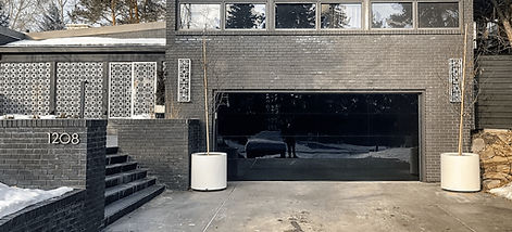 Modern mid-century style home with dark brick exterior, large upper windows, and a sleek black full-view glass garage door, surrounded by mature trees and a clean concrete driveway.