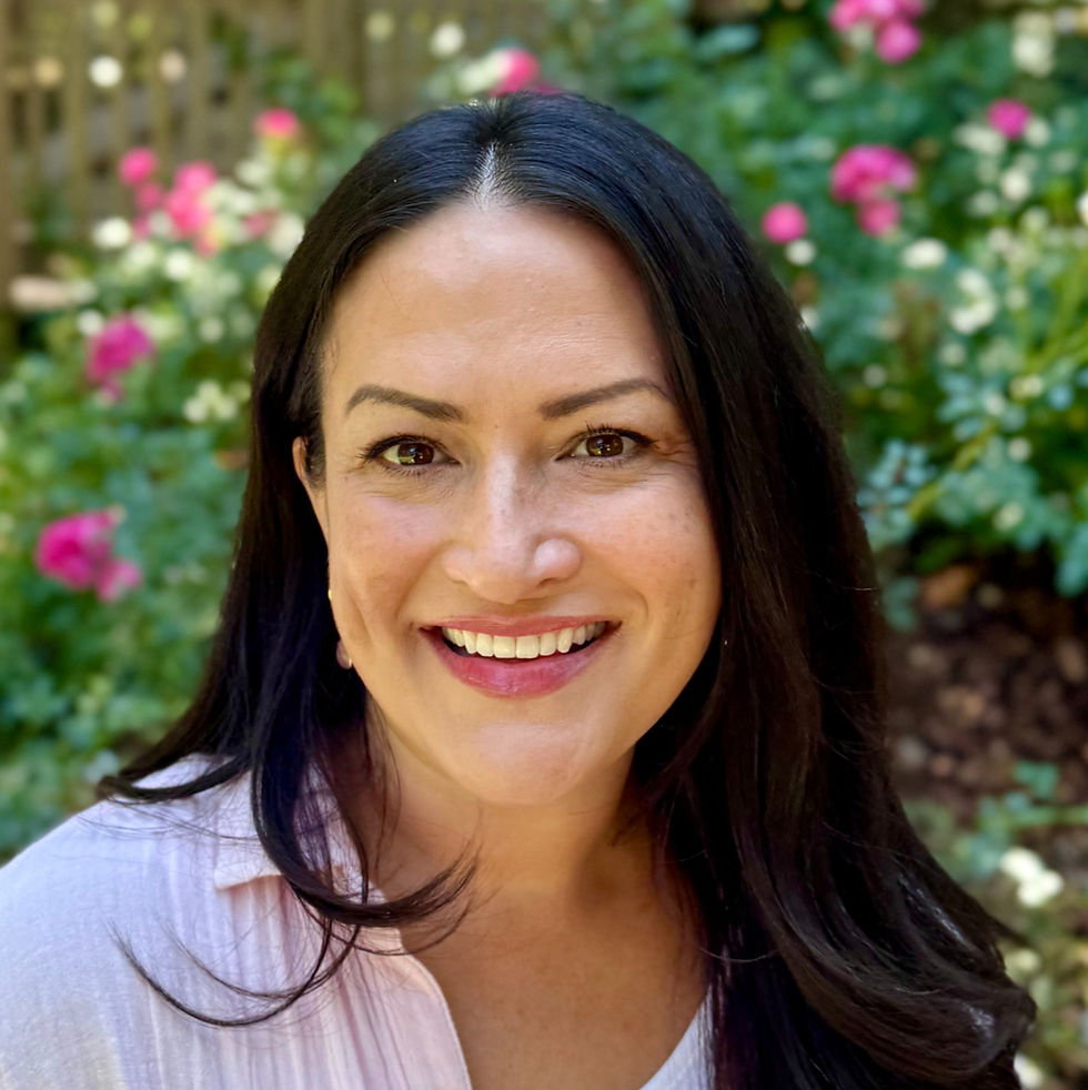 Smiling woman with long dark hair in a garden setting, surrounded by vibrant pink flowers. Bright, cheerful mood.  Jen Terrell Therapist