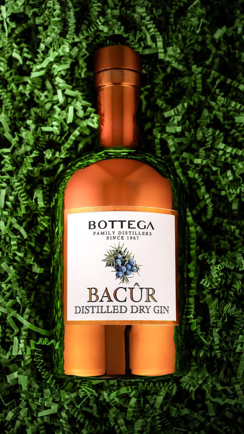 Bouteille de gin rose Bottega Bacûr sur papier vert froissé, étiquette blanche lisible, ambiance élégante.