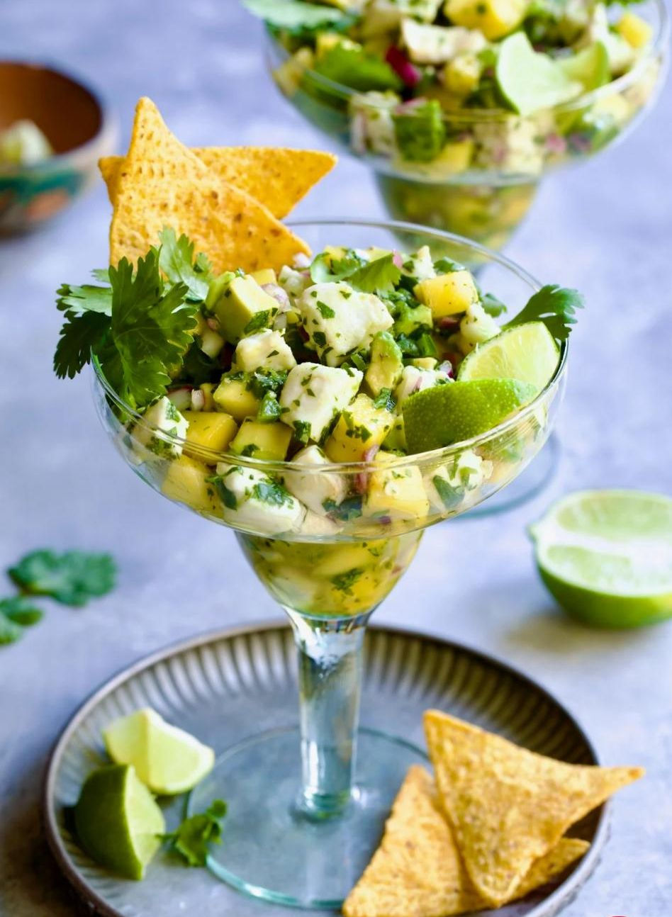 Halibut ceviche in a glass, garnished with lime, cilantro, and tortilla chips. Set on a gray surface with a festive, fresh vibe.