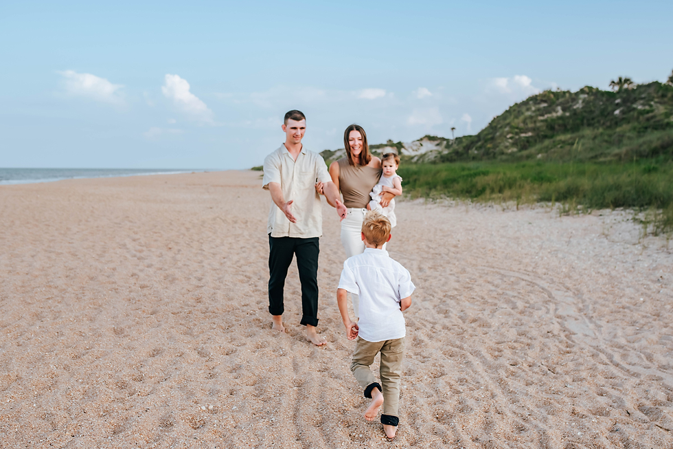 son runs to dad while mother holds baby sister on grassy sun beach for family photos