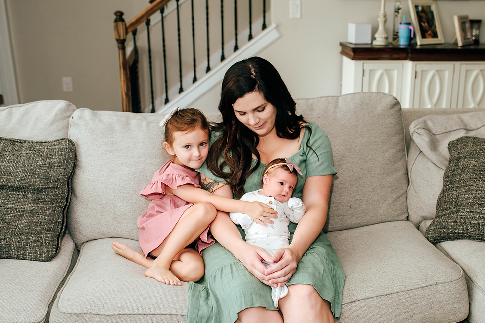 mother holds newborn baby girl in her lap on her couch as older daughter sits beside her