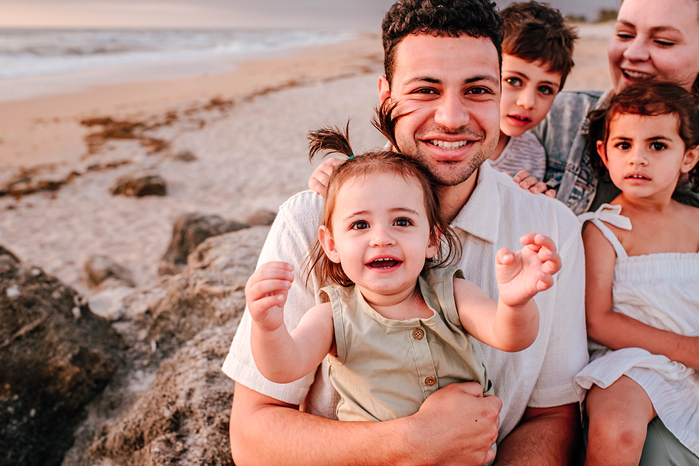 Family sitting on a beach, smiling at sunset. A man holds a laughing baby in a green dress. Kids and another adult in the background.