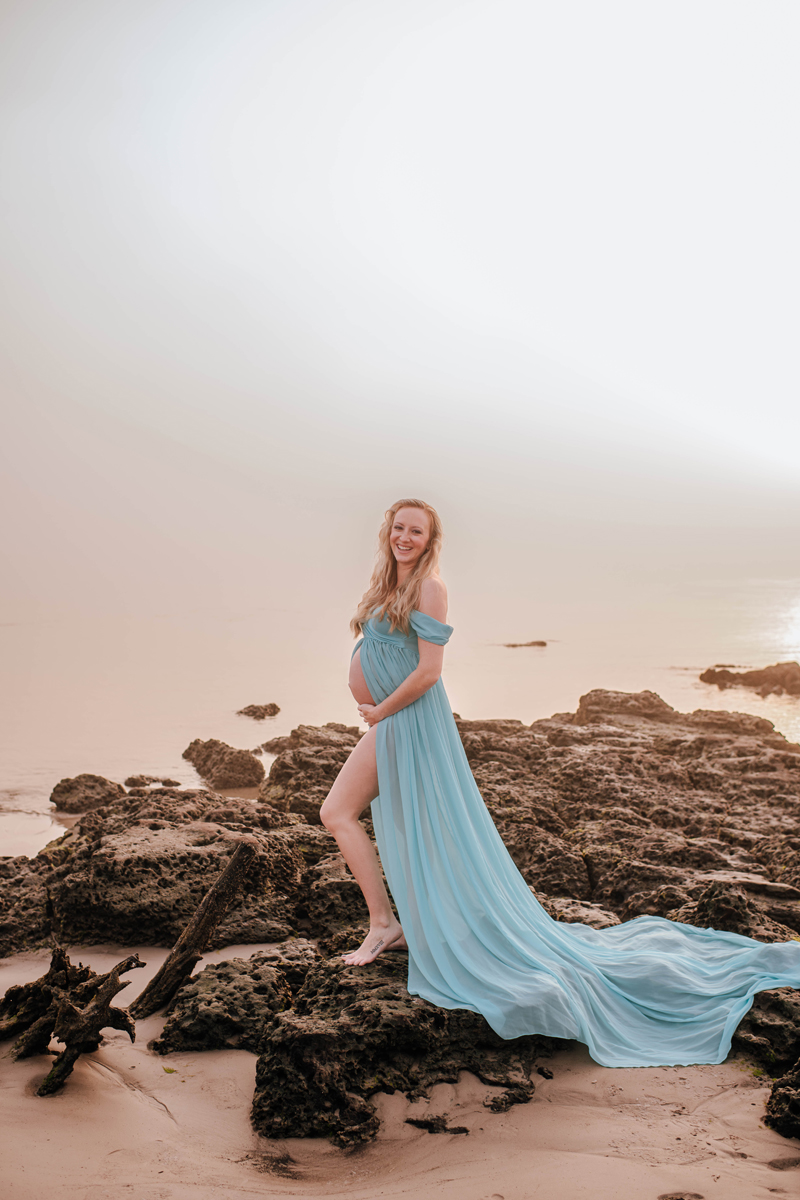 Pregnant woman in a flowing blue dress stands on rocky beach at sunset, smiling. Sky is hazy, with soft light reflecting on water.