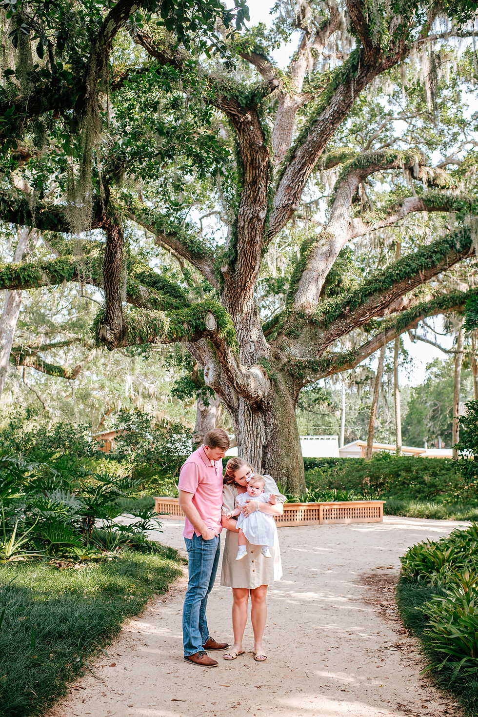 Family standing under a large tree in a park, parents holding a baby. Bright day, lush greenery surrounds them. Peaceful and joyful mood during their St Augustine family photography session.