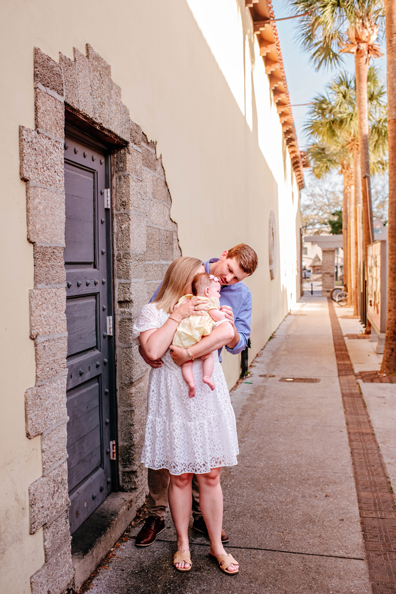 A couple embraces their baby in a yellow dress, standing near a stone wall and black door. Palm trees line the sunlit alleyway.