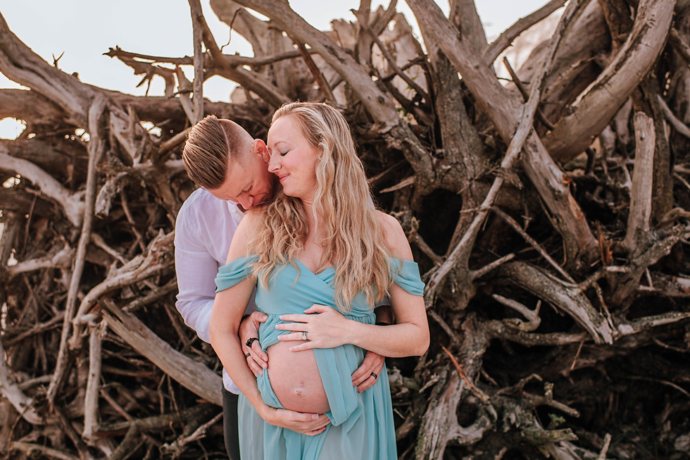 Pregnant couple embracing tenderly in front of driftwood. Woman in a light blue dress, man in white shirt. Warm, serene mood.