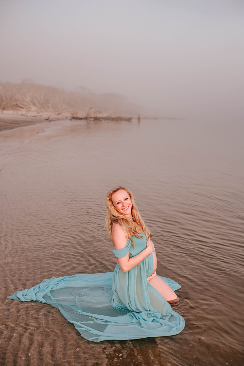 Pregnant woman kneels in shallow water, wearing a flowing teal dress. She smiles warmly, with a misty beach and bare trees behind her during her maternity photoshoot.