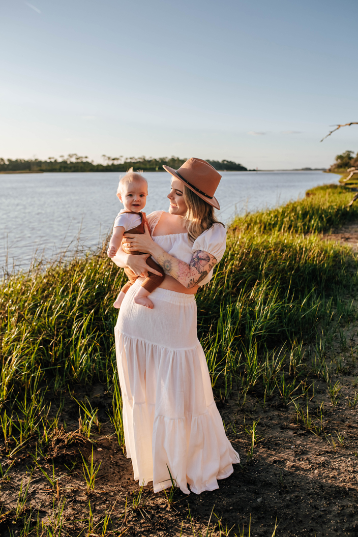 mother in white dress holding baby boy in overalls near river during family photoshoot
