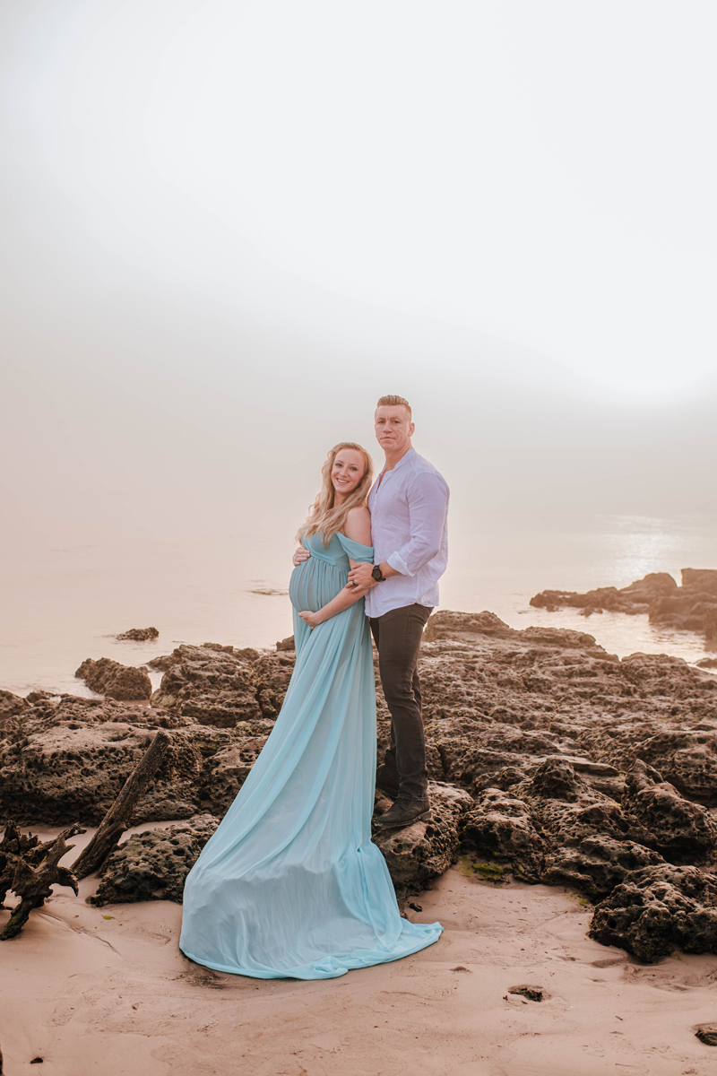 Pregnant woman in a flowing blue dress stands on rocky beach at sunset, embraced by a man in a light shirt. Serene and loving mood.