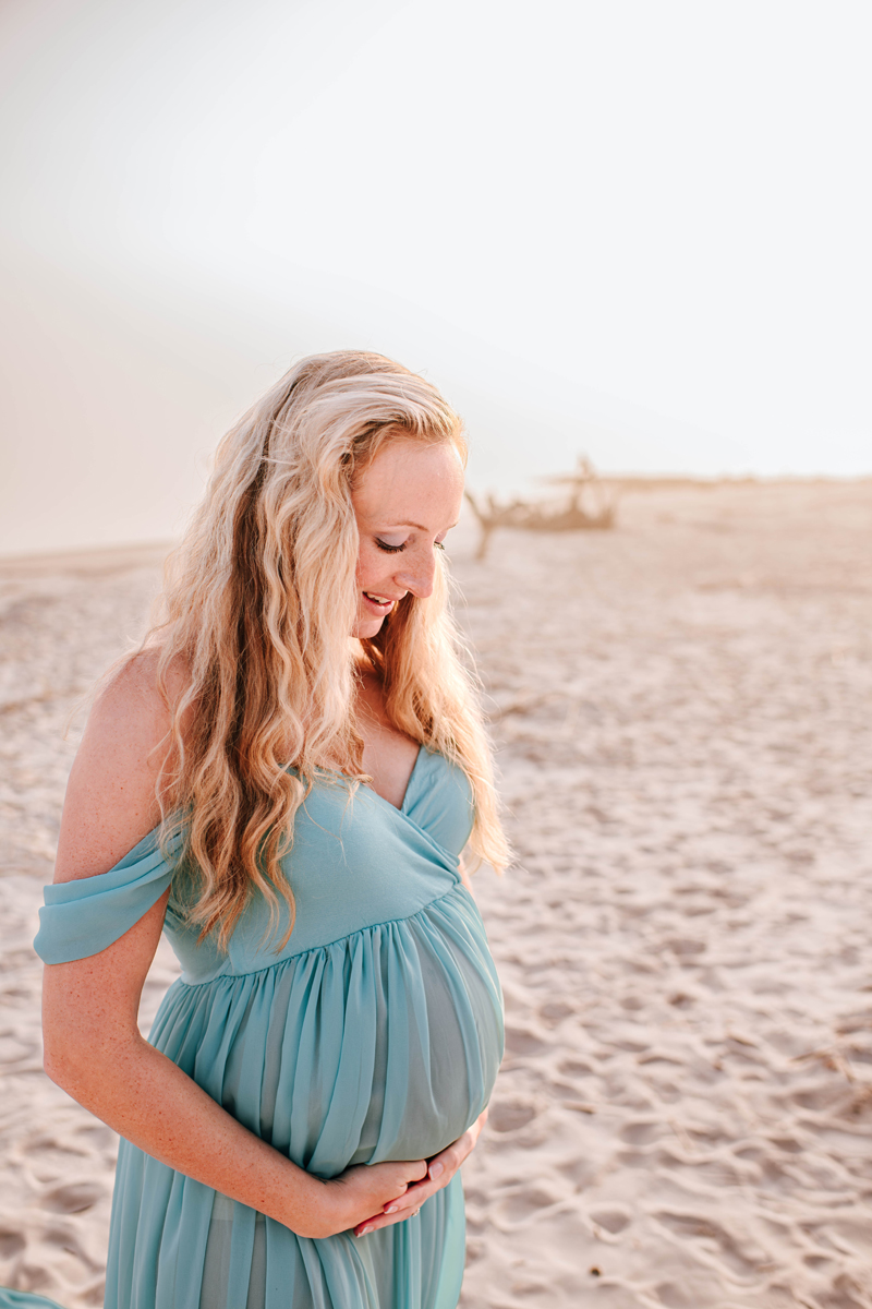 Pregnant woman in a flowing teal dress stands on a sandy beach, holding her belly with a serene smile. Soft sunlight enhances the calm mood for maternity photos.