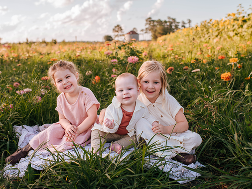 Three children smiling, sitting on a blanket in a field of wildflowers. The sky is partly cloudy, and colors are vibrant and warm.