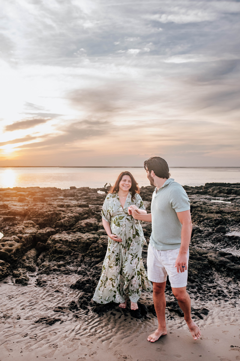husband helps his pregnant wife off the rocks on the beach with the sunrise over the ocean behind them during a maternity photoshoot