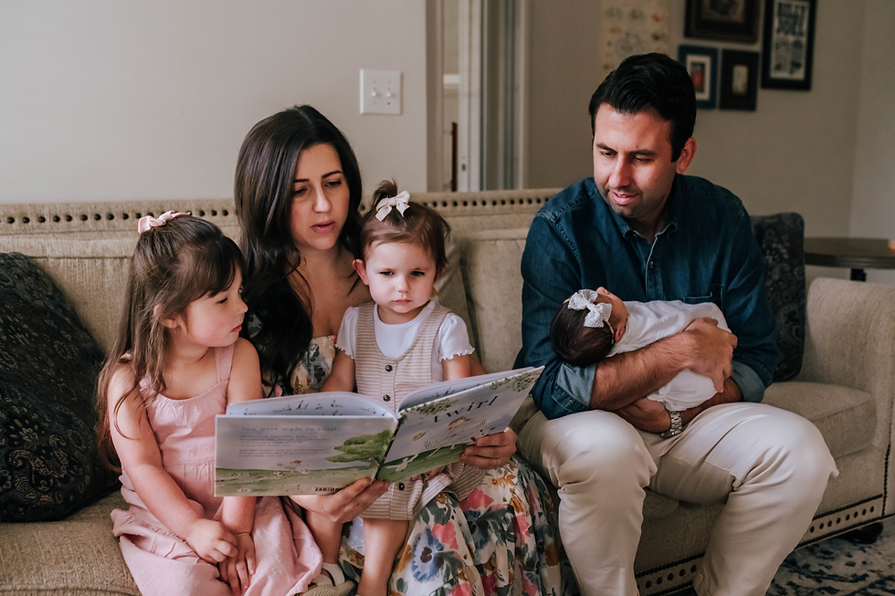 mother reads book to older daughters while dad holds newborn and looks on