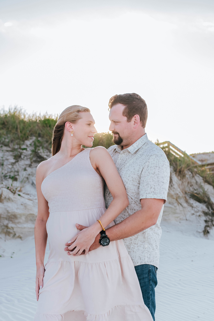 expecting couple looks at each other with sunset have during beach maternity portrait session