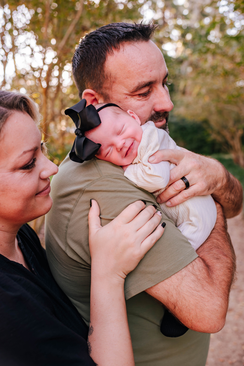 A couple outdoors lovingly holds a sleeping baby with a black bow. Warm sunlight filters through trees, creating a serene, joyful mood.