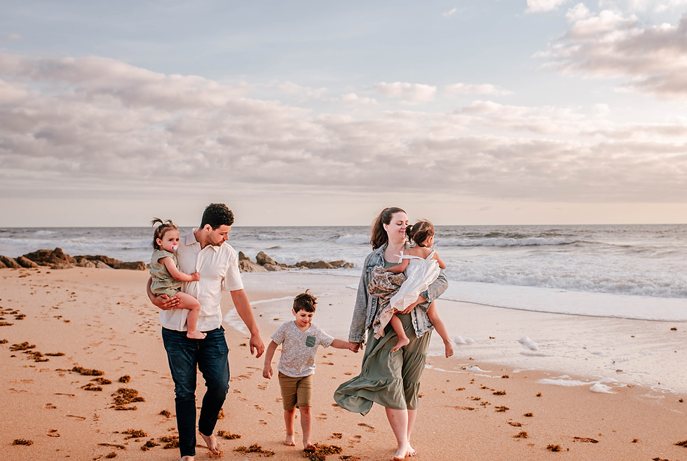 Family of five walks on a sandy beach at sunset, parents holding two children. Waves and rocks in background, creating a serene atmosphere for family pictures.