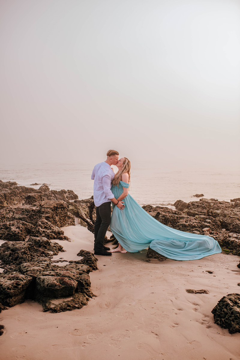 A couple kisses on a rocky beach at sunset. The woman in a flowing turquoise dress, creating a romantic, serene atmosphere for their maternity pictures.