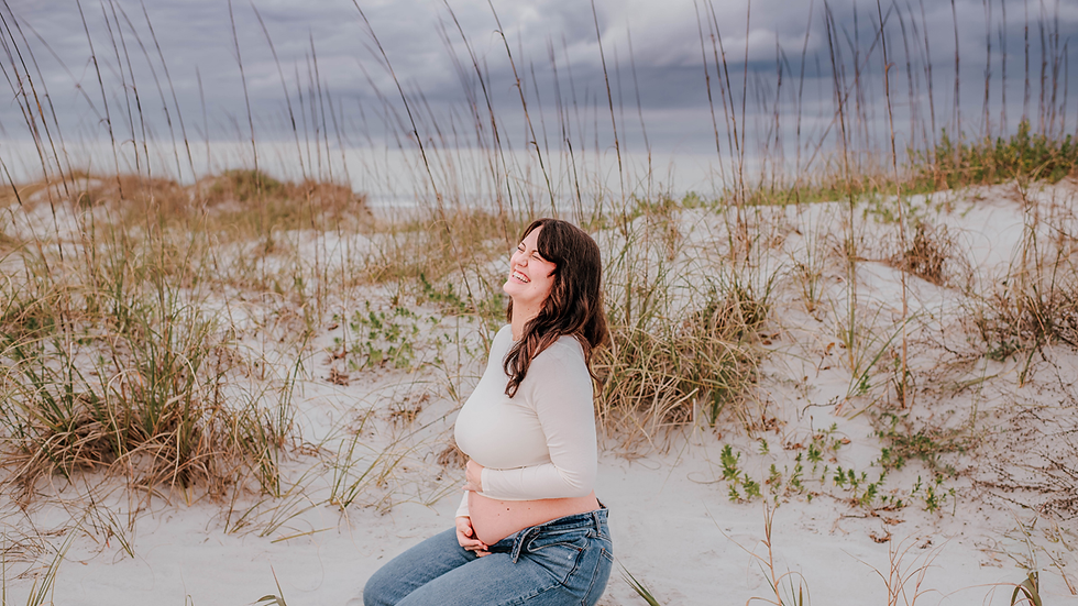 Pregnant woman kneeling on sandy beach, smiling with eyes closed. Overcast sky, tall grass, and dunes in the background create a serene mood.