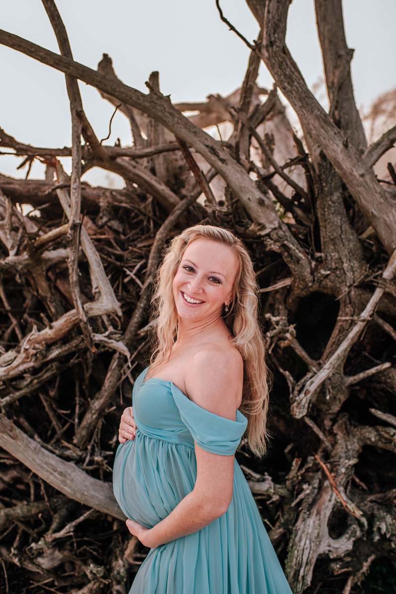 Pregnant woman in a flowing teal dress smiles, standing against a backdrop of twisted tree roots. Bright, natural setting.