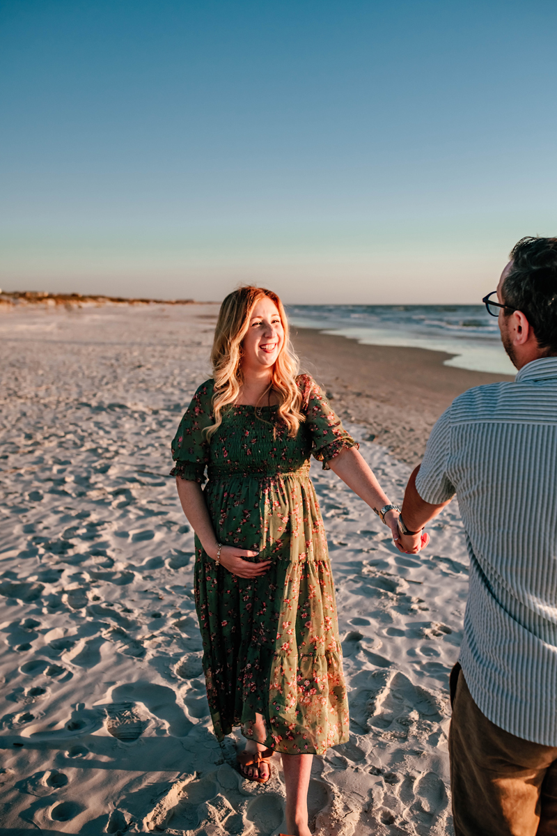 expectant mother holds hands with dad leading as mother smiles at him walking on the beach during a beach maternity photoshoot