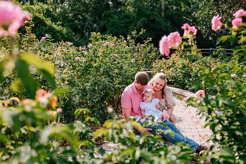 Family in garden sits on path surrounded by pink roses. Parents, smiling, hold a baby in white. Greenery creates a serene, joyful setting for family photos in St Augustine Florida.