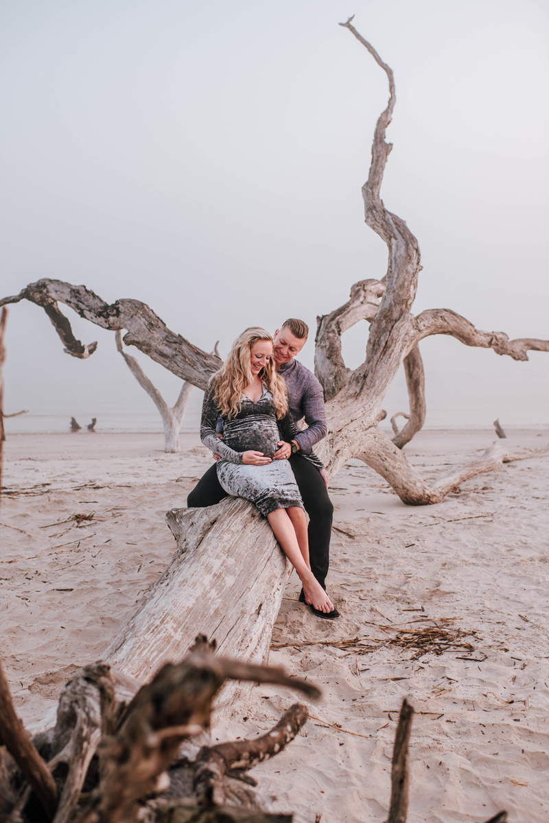 A couple sits on a driftwood log at a beach, woman holding her belly, man embracing her. The scene is serene with pale sand and sky.