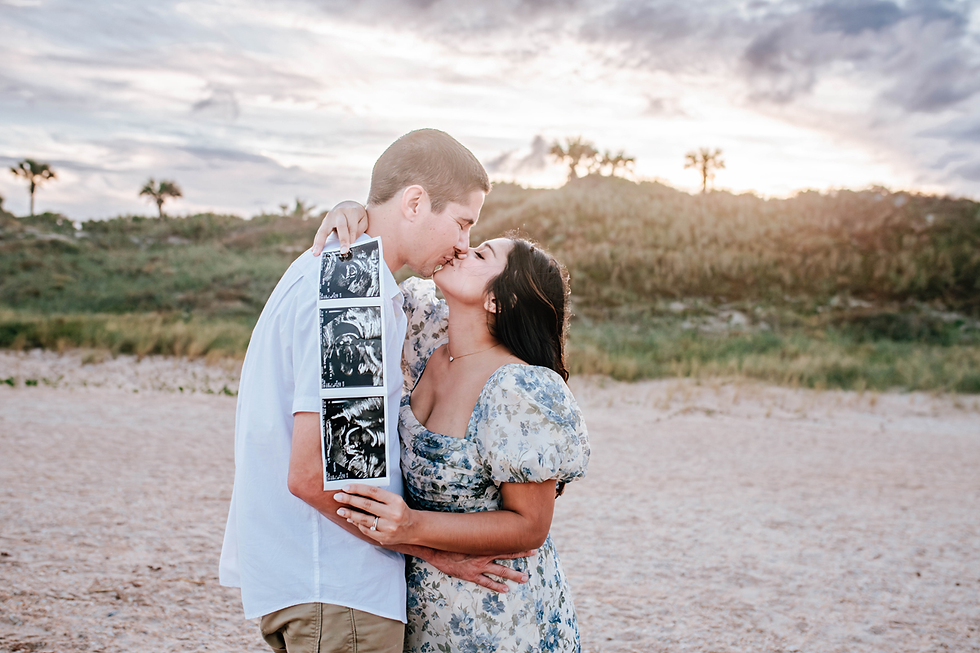 Couple kissing on a sandy beach holds ultrasound photos. The woman wears a floral dress. Green hills and sunset sky in the background. Romantic mood.