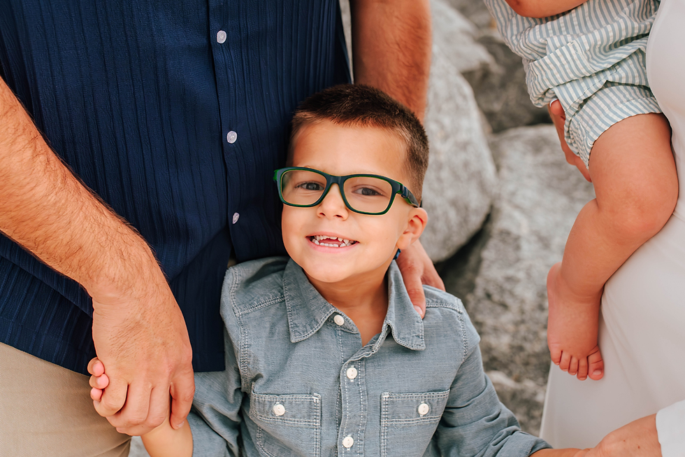 A smiling child with glasses, wearing a denim shirt, holds an adult's hand. Another adult holds a baby. Background has large rocks.